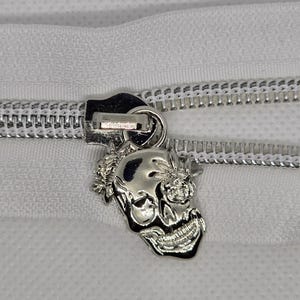 May include: Close-up of a silver zipper pull with a skull design. The skull has detailed features and is attached to a white zipper. The zipper is on a white fabric background. The zipper pull is a decorative element.