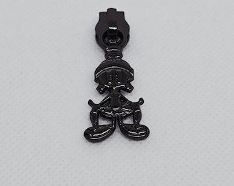 May include: A black metal zipper pull featuring a cartoon character design. The pull has a rectangular top and a detailed figure of a character with a hat, arms, and legs. The zipper pull is designed for use in clothing or accessories.