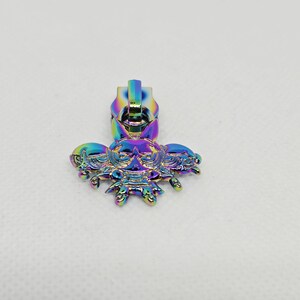 May include: A metal zipper pull featuring an iridescent rainbow finish. The pull is shaped like a stylized creature with large eyes, pointed ears, and multiple small appendages extending from the bottom, displaying vibrant blue, purple, and green hues.