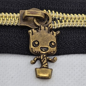 May include: A bronze-colored zipper pull featuring a stylized character design. The zipper pull is attached to a black zipper with gold-colored teeth. The character has a smiling face and is in a pot.