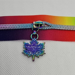 May include: A zipper tape featuring a vibrant rainbow gradient of red, orange, yellow, green, blue, and purple. Attached to the zipper slider is a metallic, iridescent maple leaf charm with an intricate, embossed filigree pattern throughout.
