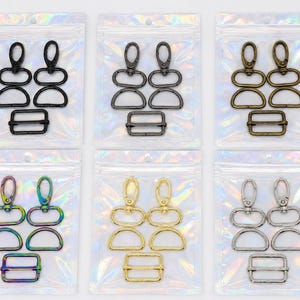 May include: Six sets of metal hardware for bags, each in a clear iridescent pouch. Each set contains two swivel lobster clasps, two D-rings, and one rectangular slider buckle. The hardware comes in black, gunmetal, bronze, rainbow, gold, and silver.