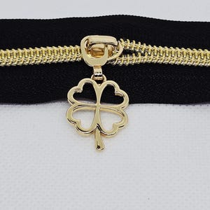May include: Close-up of a black zipper with gold-colored teeth and a gold-colored four-leaf clover charm. The zipper is on a white background.