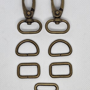 May include: An assortment of antique brass-colored hardware components, including two swivel clasps, two D-rings, two rectangular rings, and a double bar slider. These metal findings are arranged on a white background.