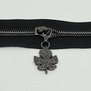 May include: A black zipper featuring a metallic gunmetal slider pull shaped like a detailed maple leaf. The zipper tape is black with matching black teeth, and the leaf-shaped charm is attached to the slider by a small circular jump ring.