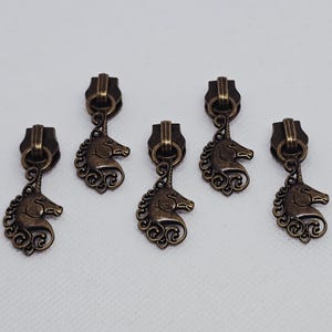 May include: Five antique bronze zipper pulls with a unicorn design. Each pull features a rectangular top with a loop and a detailed unicorn head pendant with a flowing mane and a spiraling design.