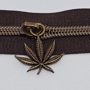 May include: A brown zipper with a bronze-colored pull tab and teeth. The pull tab is a cannabis leaf charm. The zipper is set against a neutral background.