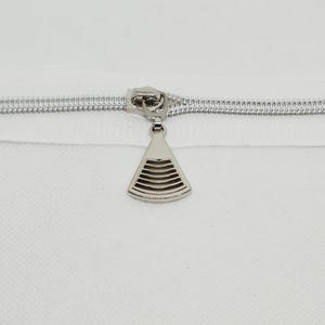 May include: A silver-toned metal zipper pull attached to a white zipper tape. The pull features a fan-shaped design with horizontal cutouts, creating a series of parallel lines within the metallic frame, centered on the zipper track.