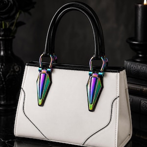 May include: A structured white handbag featuring black top handles and iridescent, rainbow-colored metallic hardware accents. The bag has a clean, rectangular silhouette with black piping details and two prominent, pointed metallic attachments.