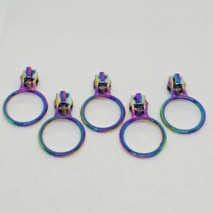 May include: Five metal zipper pulls with a circular ring design and an iridescent rainbow finish. Each pull features a polished, multi-colored metallic surface that shifts between shades of blue, purple, green, and yellow, arranged in a row.