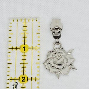 May include: A silver-toned metal zipper pull featuring a detailed rose-shaped charm. The zipper pull is positioned next to a clear plastic ruler, showing the charm measures approximately 1 inch in width and height, excluding the pull mechanism.