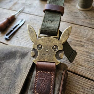 May include: An olive green canvas bag featuring a brass-toned metal buckle shaped like the head of a cartoon character with pointed ears. The buckle is attached to a matching olive green canvas strap and a dark brown textured leather tab with stitching.