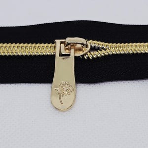 May include: Close-up of a black zipper with gold-colored teeth and a gold pull tab. The pull tab features a rose design. The zipper is on a white background.