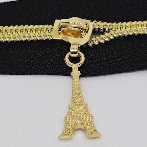 May include: A gold-colored zipper with a black fabric backing. The zipper pull features a detailed Eiffel Tower charm. The zipper teeth and pull are a bright gold color, and the charm is intricately designed.