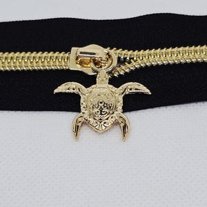 May include: Close-up of a gold-colored zipper with a black fabric backing. The zipper pull is a detailed turtle pendant. The zipper teeth are also gold, creating a contrast with the black fabric.