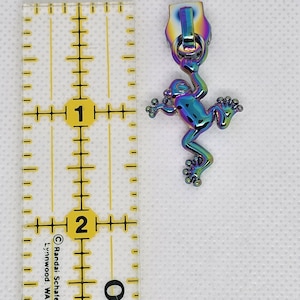 May include: Iridescent zipper pull with a frog design. The frog is approximately 1 inch in height. The zipper pull has a rainbow finish, with a matching pull tab. The background is white.