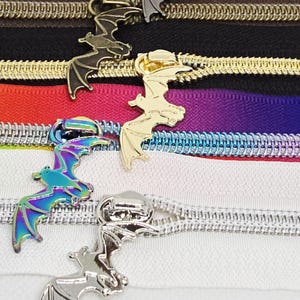 May include: A collection of five zipper tapes featuring bat-shaped metal pulls. The zippers include black, brown, gold, iridescent rainbow, and silver finishes, each paired with matching or contrasting fabric tape in black, brown, purple, and white.