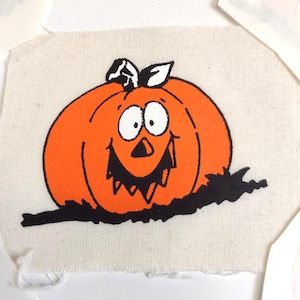 May include: A square piece of off-white fabric featuring a screen-printed illustration of a bright orange pumpkin with a black outline. The pumpkin has a wide, jagged black smile, large white eyes, and a black stem, resting on a black patch of ground.