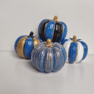 May include: A collection of four decorative pumpkins featuring distinct finishes. One is blue and black with a gold stem, one is blue and gold, one is textured blue and metallic bronze, and one is blue and white with a gold stem.