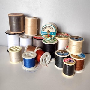 May include: A collection of vintage thread spools in various colors, including white, black, brown, blue, and beige. Some spools have labels with text, such as "Fruit of the Loom" and "Lily". The spools are made of wood and plastic.