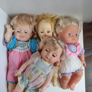 May include: Four plastic baby dolls with blonde hair and painted facial features. One doll wears a blue and pink outfit, another a white romper with purple hearts, and a third a pastel patterned dress. The dolls have articulated limbs and eyes.