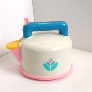 May include: A vintage Fisher-Price toy watering can in white, blue, pink, and yellow. The watering can has a blue handle, a pink spout, and a pink base. The logo of a flower is on the front.
