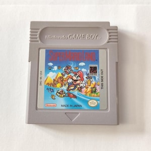May include: A gray Nintendo Game Boy cartridge featuring the game "Super Mario Land." The label shows Mario in a colorful action scene. The cartridge has the text "Nintendo GAME BOY" and "MADE IN JAPAN."