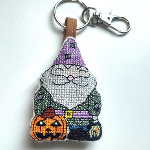 May include: A cross-stitched keychain featuring a gnome with a purple hat, a gray beard, and a green tunic. The gnome holds an orange jack-o'-lantern pumpkin in one hand and a small lantern in the other, attached to a silver metal keyring.