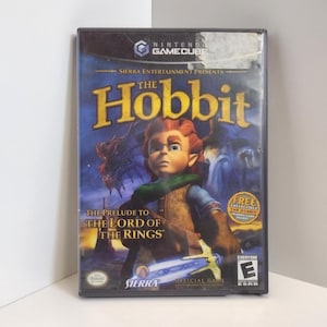 May include: A Nintendo GameCube video game case for "The Hobbit." The cover art features an illustration of a character with red hair and a green scarf, with the title in large gold letters. The game is rated "E" for Everyone.