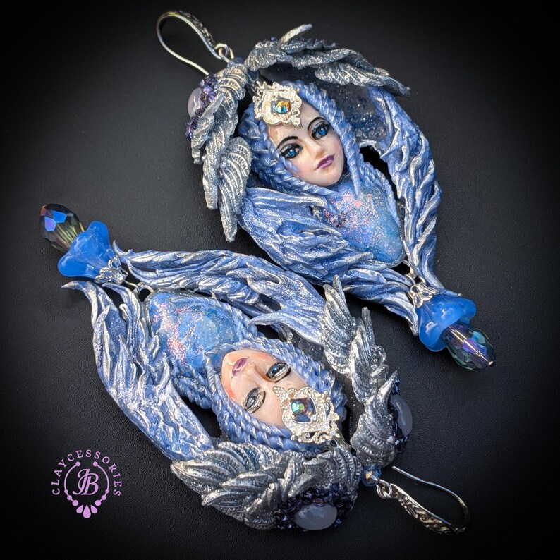 May include: A pair of handcrafted earrings featuring a blue-haired woman's face with a silver crown and accents. The earrings have a detailed, sculpted design with blue and silver tones, and a dangling blue crystal. The earrings are set against a black background.
