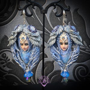 May include: A pair of ornate, fantasy-themed earrings. Each earring features a sculpted face with blue hair and a silver-colored headdress. The design includes blue and silver wings and a dangling, iridescent crystal. The earrings are displayed on a black stand.