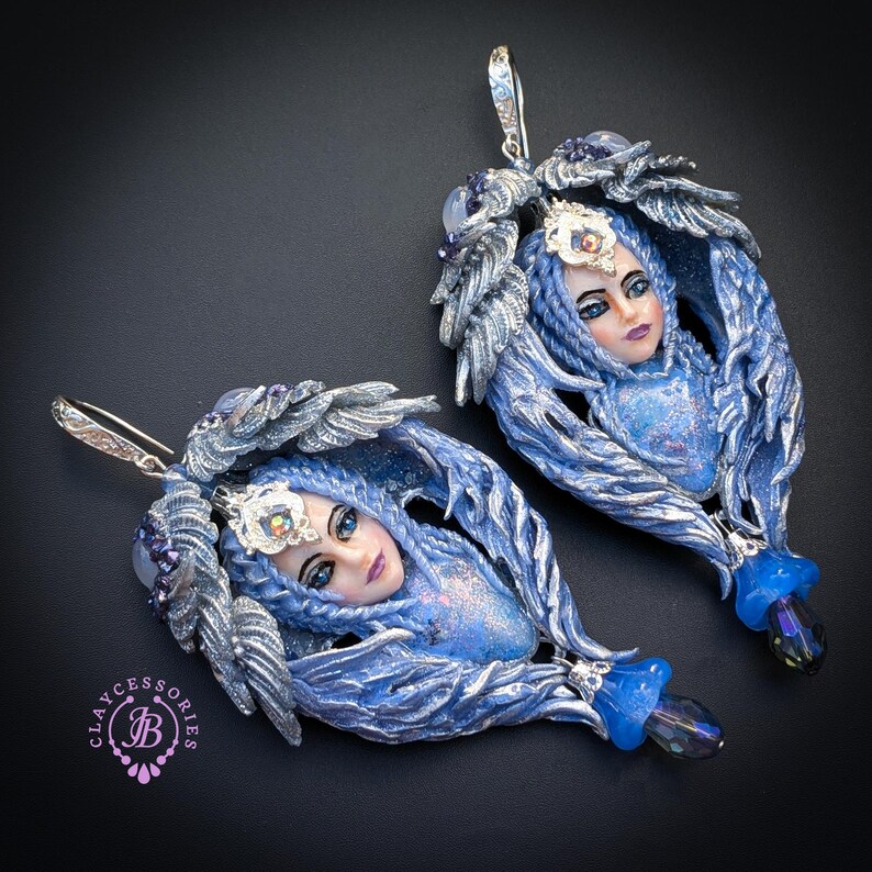 May include: A pair of handcrafted blue and silver earrings. Each earring features a sculpted face with blue hair, a silver crown, and is surrounded by silver-colored feather-like details. The earrings have a blue teardrop-shaped bead at the bottom.