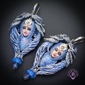 May include: A pair of handcrafted blue and silver earrings. Each earring features a sculpted face with blue eyes and a silver crown, surrounded by blue and silver feather-like details. The earrings have a hook closure and a dangling blue bead.