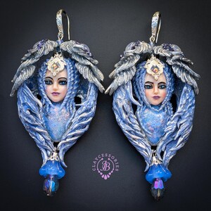 May include: A pair of handcrafted blue and silver earrings. Each earring features a sculpted face with blue hair, a jeweled crown, and a floral accent. The earrings have a dangling blue flower and a crystal bead. The earrings are designed with a fantasy theme.