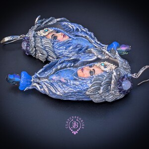 May include: A pair of handcrafted earrings featuring a woman's face with blue and silver accents. The earrings have a detailed design with a blue gemstone and a silver hook. The earrings are a unique piece of jewelry.