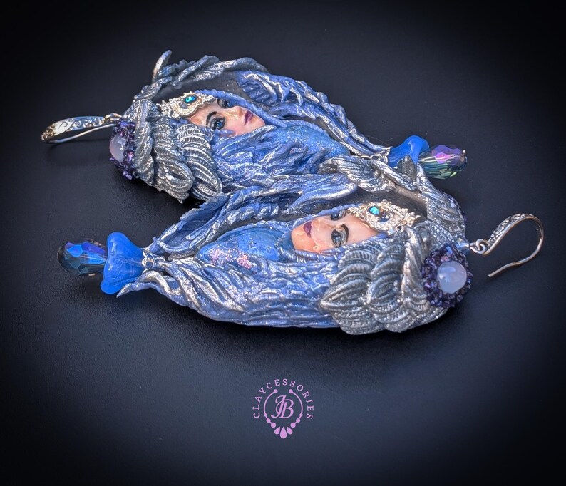 May include: A pair of handcrafted earrings featuring a woman's face with blue and silver accents. The earrings have a detailed design with a blue gemstone and a silver hook. The earrings are a unique piece of jewelry.
