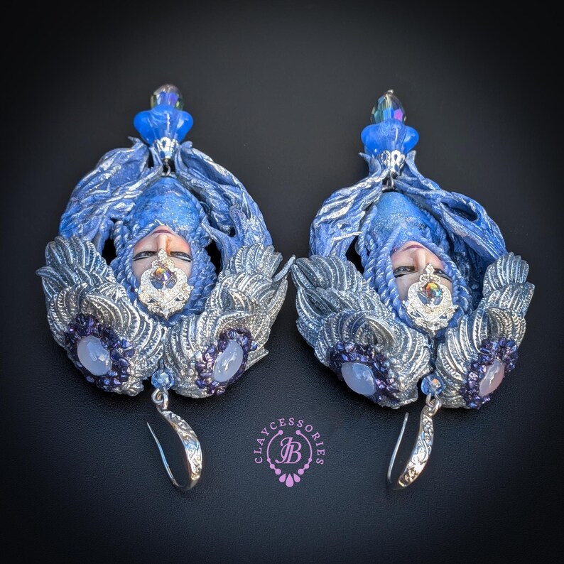 May include: A pair of ornate, handmade earrings featuring a face with blue hair and silver accents. The earrings have a dangling silver hook and are adorned with purple and clear gemstones. The design includes a crown-like embellishment.