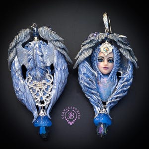 May include: A pair of fantasy-themed earrings. Each earring features a blue and silver winged design with a central face and a dangling blue crystal. The earrings have a detailed, handcrafted appearance, with intricate feather and face details.