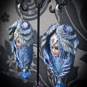 May include: A pair of ornate, blue and silver fantasy earrings. Each earring features a sculpted face with a headdress and flowing details. The earrings have a teardrop shape with a blue gem at the bottom and a silver hook.