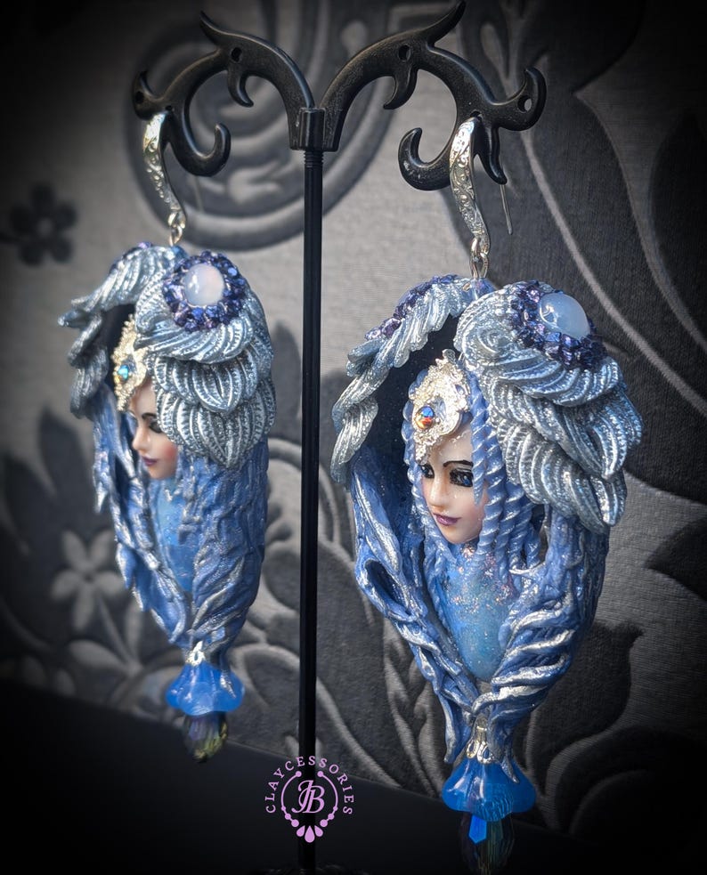 May include: A pair of ornate, blue and silver fantasy earrings. Each earring features a sculpted face with a headdress and flowing details. The earrings have a teardrop shape with a blue gem at the bottom and a silver hook.