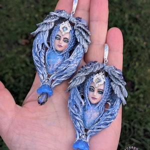 May include: A pair of blue and silver fantasy-themed earrings. Each earring features a sculpted face with a crown, surrounded by feathery wings. A blue gem hangs from the bottom, and a silver hook is at the top.