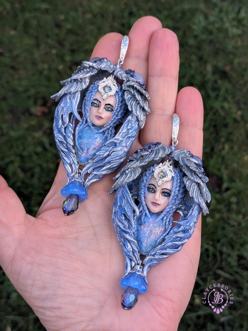 May include: A pair of blue and silver fantasy-themed earrings. Each earring features a sculpted face with a crown, surrounded by feathery wings. A blue gem hangs from the bottom, and a silver hook is at the top.