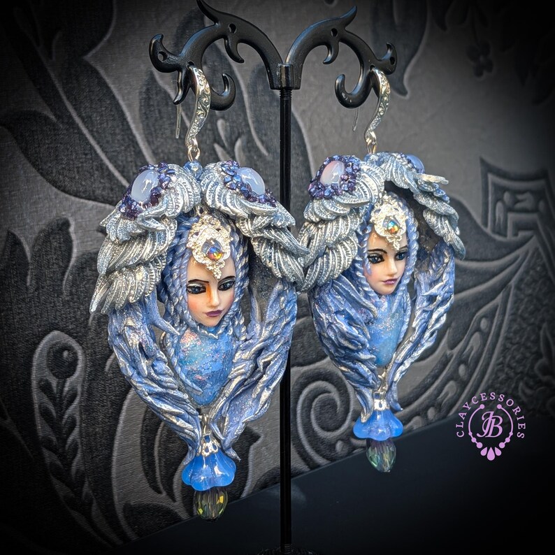 May include: A pair of ornate, blue and silver earrings featuring sculpted faces with detailed features. The earrings have a fantasy theme, with intricate feather-like designs and sparkling accents. The earrings are displayed on a black stand.