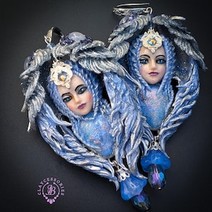 May include: A pair of heart-shaped pendants featuring detailed fairy faces with blue hair and silver accents. Each pendant has a blue and silver wing design, adorned with a jeweled crown and dangling blue flower charms. The pendants are set against a dark background.