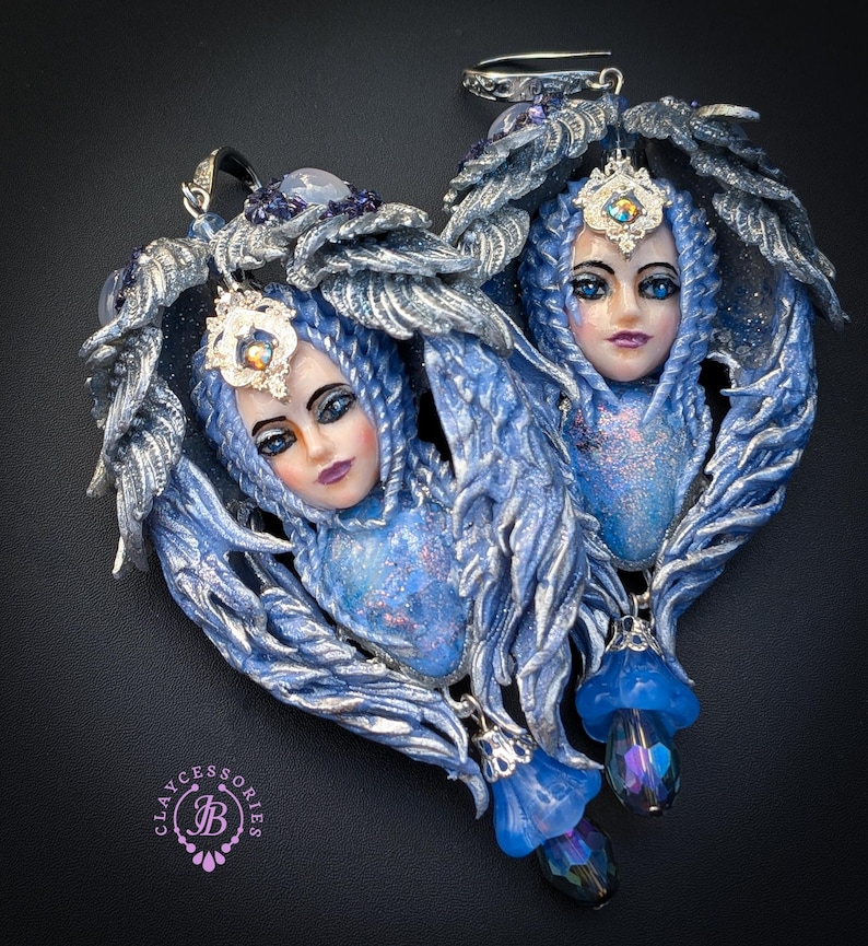 May include: A pair of heart-shaped pendants featuring detailed fairy faces with blue hair and silver accents. Each pendant has a blue and silver wing design, adorned with a jeweled crown and dangling blue flower charms. The pendants are set against a dark background.