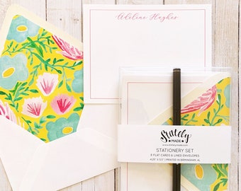 May include: A set of stationery featuring a floral design with pink, blue, and yellow flowers on a yellow background. The set includes 8 flat cards and lined envelopes, measuring 4.25 inches by 5.5 inches. The stationery is printed in Birmingham, Alabama.