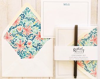 May include: Stationery set with white cards and envelopes. The envelopes have a floral pattern lining with pink and blue flowers. A black pen rests on the set, which includes a notepad with the initials "MLG". The set is packaged in a clear box.
