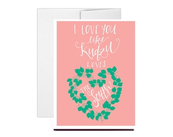 May include: A pink card with the text "I love you like Kudzu loves the South" in white lettering. A green vine in the shape of a heart is behind the text.