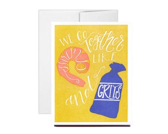 May include: A yellow card with the text "We go together like shrimp and grits" in white script. A pink shrimp and a blue bag of grits are illustrated on the card.