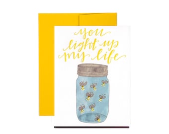 May include: A greeting card with a blue mason jar filled with fireflies and the text "you light up my life" in yellow calligraphy.